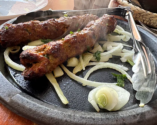 Two beef seekh kababs on a sizzling iron plate with onion rings and serving tongs