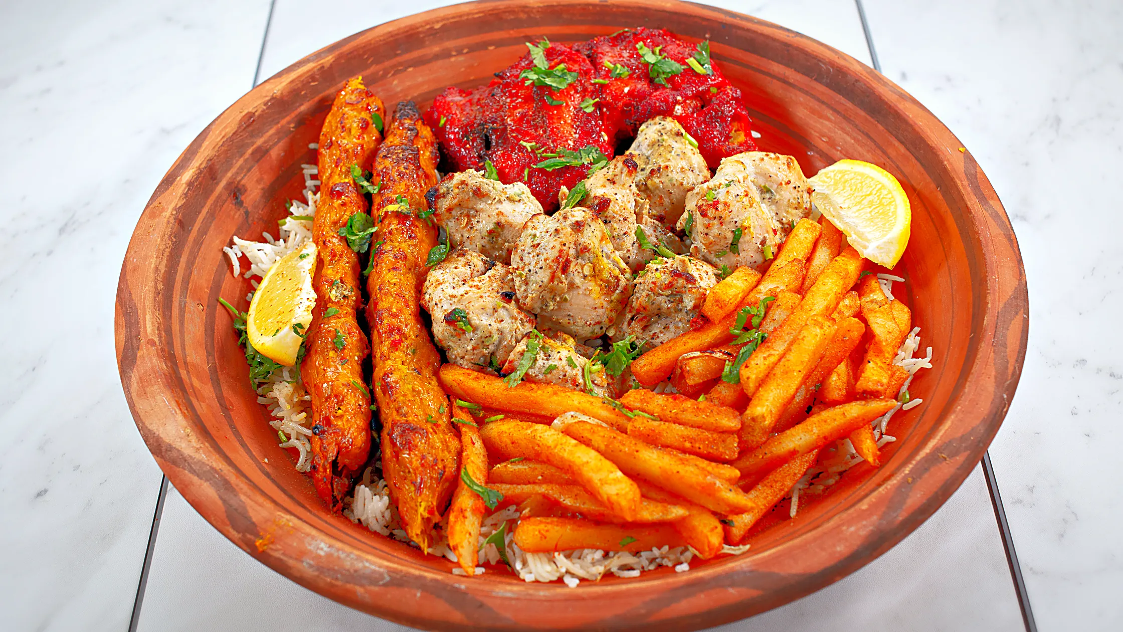 Clay bowl of mixed BBQ on rice: chicken seekh kababs, red chicken tikka, malai tikka, spiced fries and lemon wedges