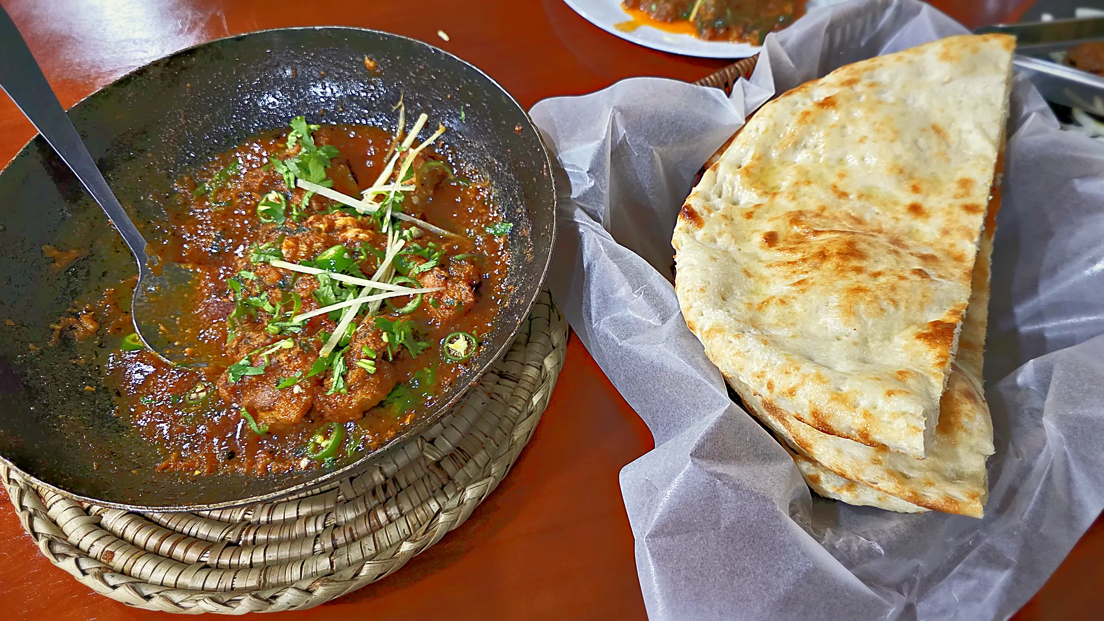 Karahi in a black iron wok on a woven mat, garnished with ginger strips and green chilies, with naan in a paper-lined basket